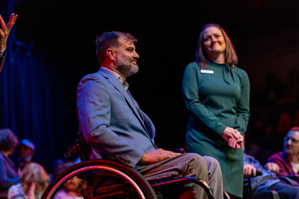 Two people on stage under warm lighting at CAN Talks, one seated in a wheelchair and one standing, both smiling toward the audience