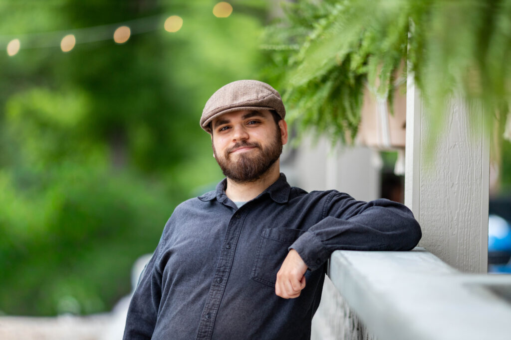 Holden Roberts leans on a porch railing with a closed-mouth smile. He has a full dark beard and wears a brown herringbone flat cap with a charcoal button-up shirt. Ferns and string lights blur in the background.