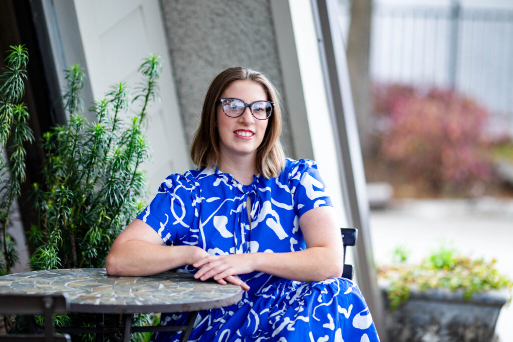 Jean Knight sits at a mosaic café table with her hands resting on top and smiles at the camera. She has shoulder-length light brown hair and tortoiseshell glasses and wears a cobalt dress with a white abstract print.