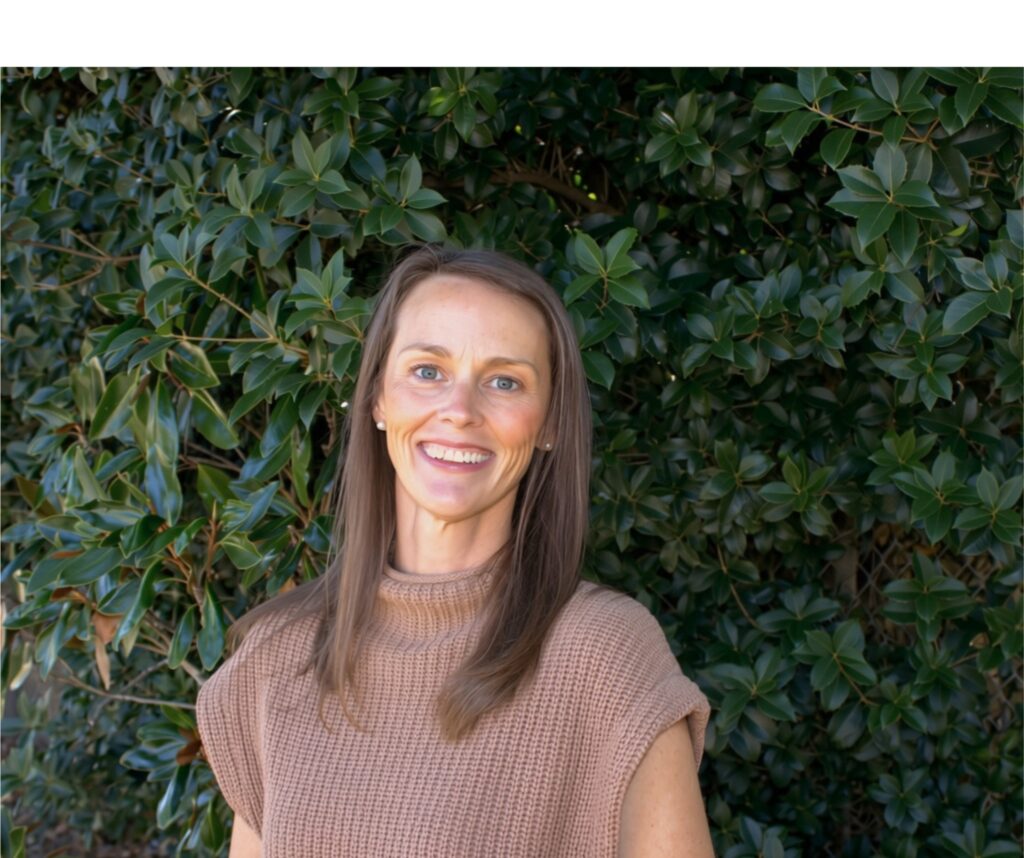 Jessie Garris smiles warmly in front of a dense wall of glossy green magnolia leaves. She has long, straight light brown hair and wears pearl stud earrings with a tan knit sweater that has a mock neck and short cap sleeves.