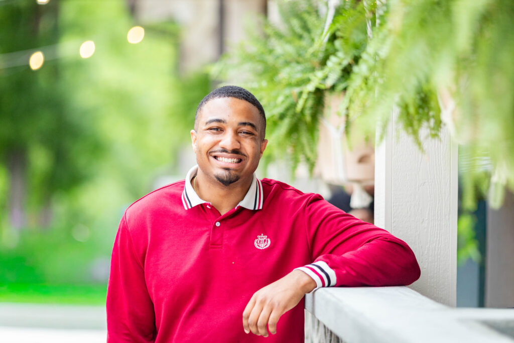Craig Garrison grins wide with one arm resting on a porch railing. He wears a red long-sleeve polo with a striped collar, and hanging ferns and string lights glow behind him.