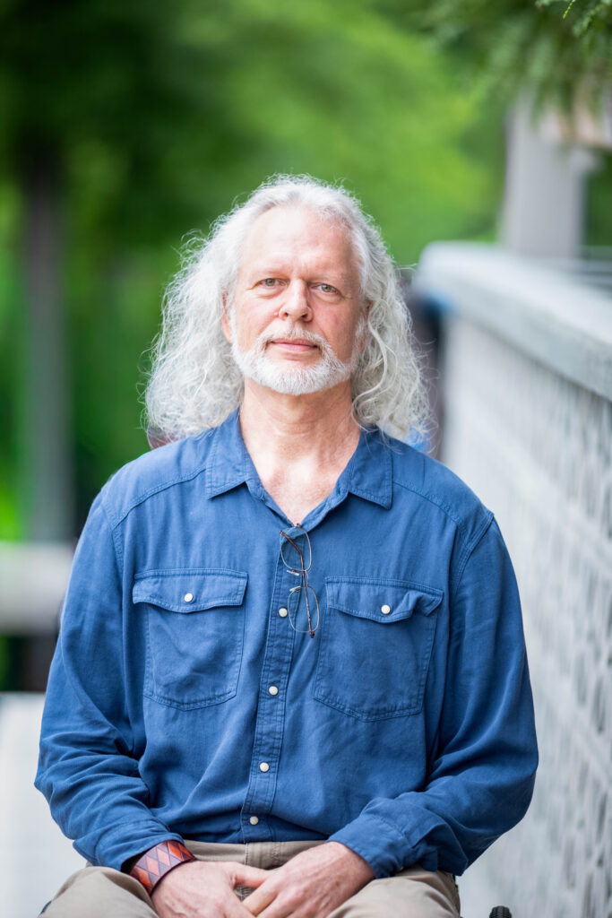 David Ackerman sits in his wheelchair beside a white breeze-block wall and looks calmly at the camera. He has long, curly silver hair and a white beard. His reading glasses hang from the front of his denim-blue shirt, and he wears a leather cuff on one wrist.