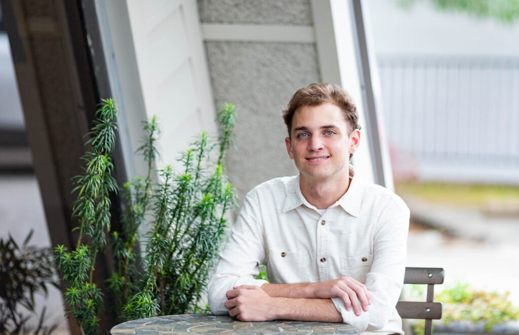 Landon Barber, a young man with wavy light brown hair, smiles at the camera. He wears a cream button-up shirt and rests his folded arms on a mosaic café table beside a tall potted plant.