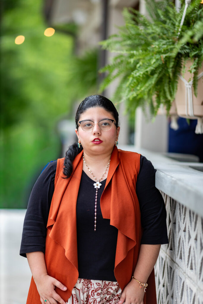 Val Hincapie stands beside a white breeze-block wall under a hanging fern. She has a long dark braid, wire-rimmed glasses and red lipstick. She's wearing a rust-orange draped vest over a black top, a long pearl necklace with a white flower pendant and a patterned skirt.