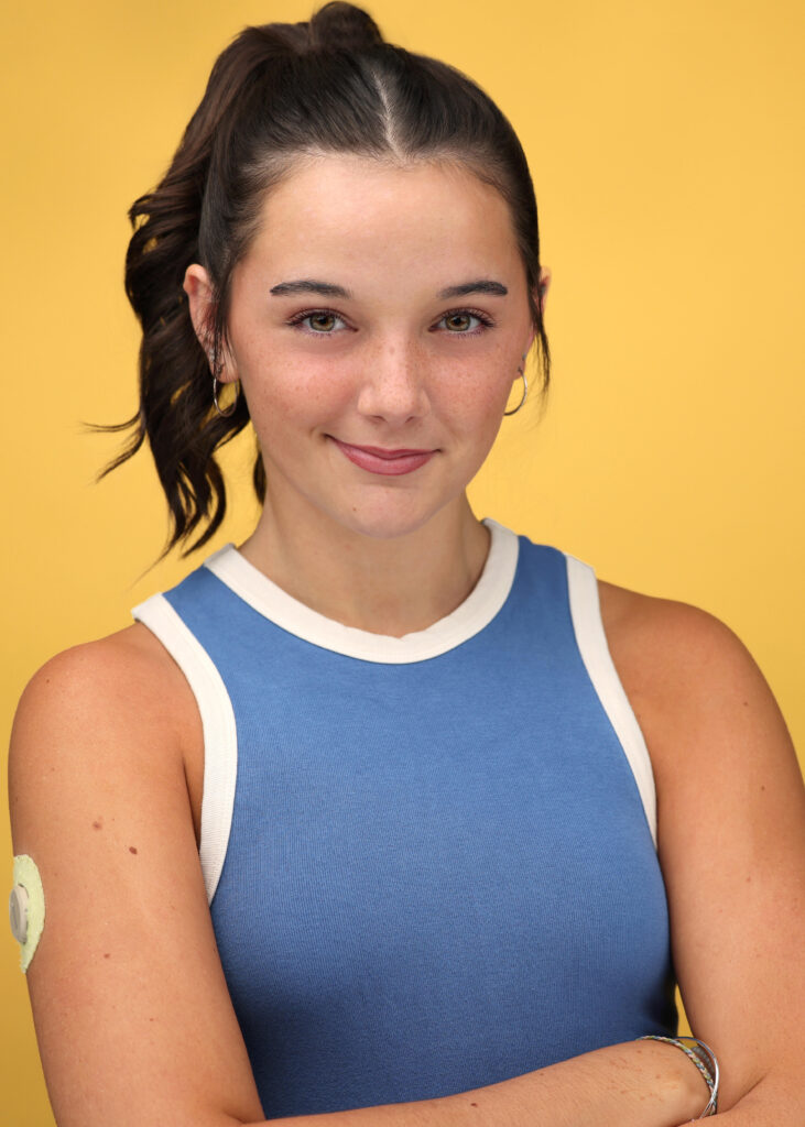 Riley Fincher-Foster stands with her arms crossed against a bright yellow backdrop and gives a confident smile. Her dark hair is pulled up in a high, wavy ponytail. She wears silver hoop earrings, a blue tank top with cream trim and a few thin bracelets. A small sensor is visible on her upper arm.